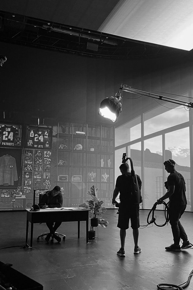 A behind-the-scenes view of a film or television production set with crew members working, a seated person at a desk, and a large window showing a cityscape outside. The background features sports memorabilia and jerseys.