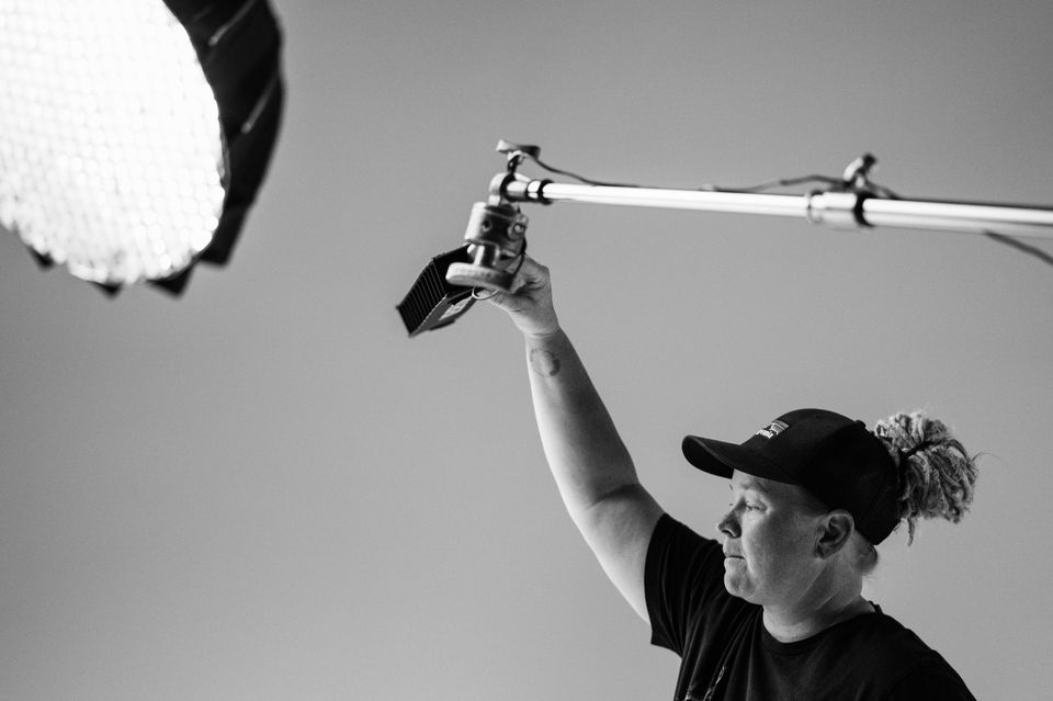 A woman wearing a cap and black t-shirt adjusting lighting equipment outdoors during daytime, with clear sky in background.