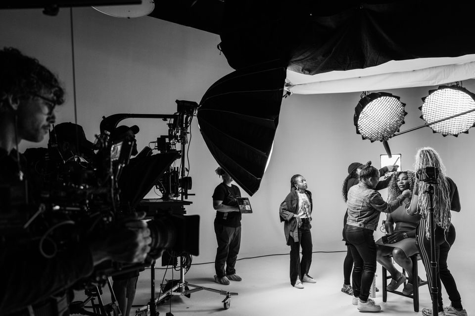 Black and white photo of a film or photo studio with crew and models. Crew members are adjusting makeup and preparing the model for filming or shooting, with professional lighting setup overhead and camera equipment visible.