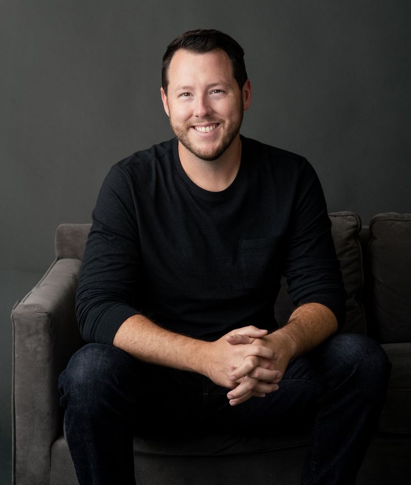 A smiling man with dark hair and a beard, wearing a black long sleeve shirt, sitting on a gray sofa with his hands clasped together. The background is gray.