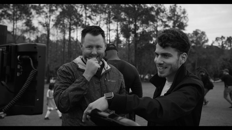 Two young men smiling and looking at a tablet outdoors, with trees and other people in the background, in a black and white photo.