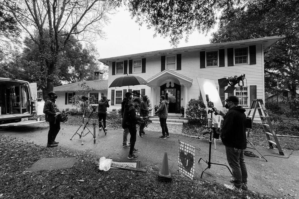 Film crew filming outside a two-story house with studio equipment and crew members, some with umbrellas, on a cloudy day.