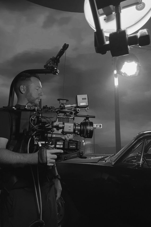 A man operating a professional film camera outdoors at dusk, with a street sign and a car visible in the background.