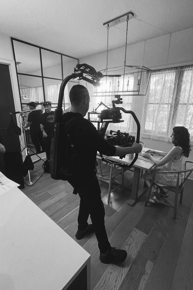 A film crew setting up shot in a dining room for a scene with a woman sitting at the table. The camera operator is focused on filming the woman, who is looking toward the camera with her hands on the table. Crew members are visible in the background 