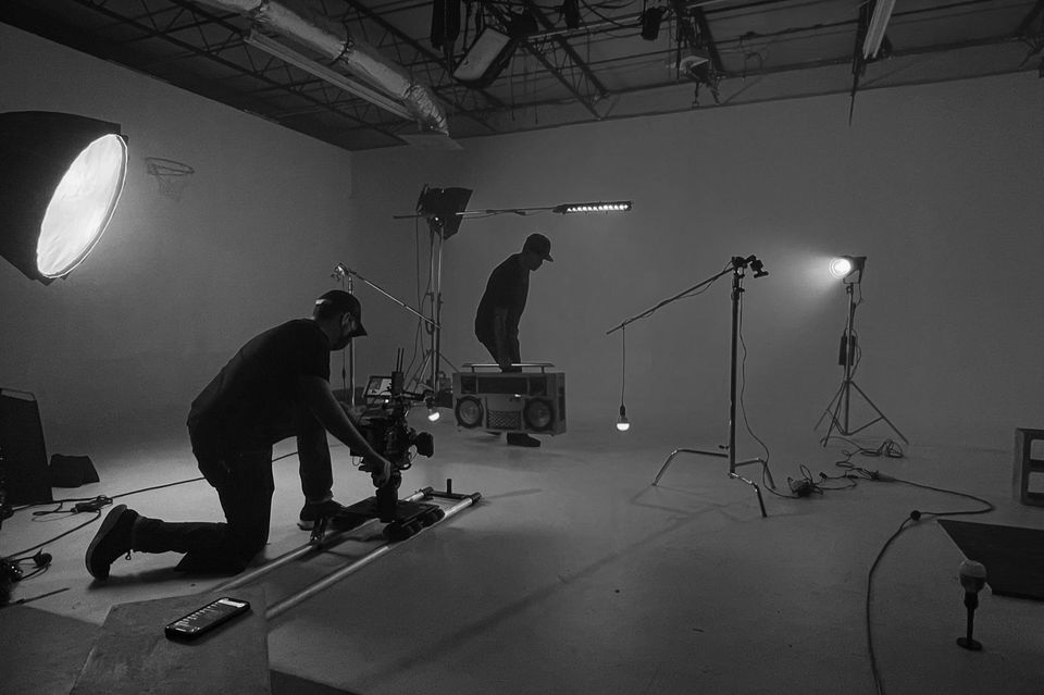 Black and white photo of a film or photo studio with two people, one kneeling and one standing, surrounded by lighting and camera equipment.