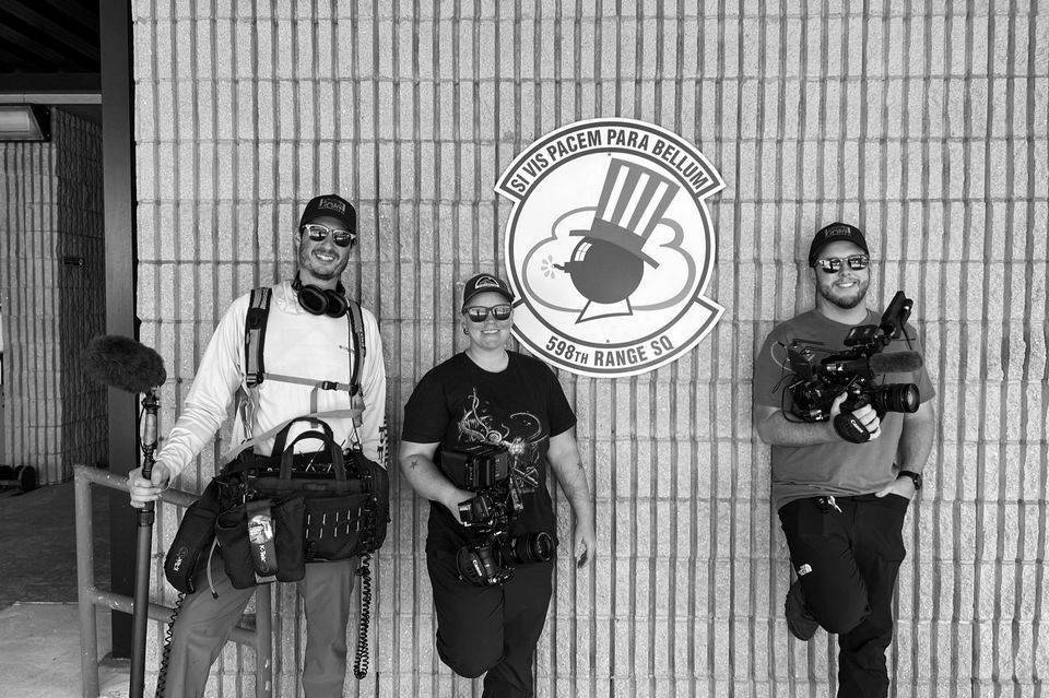 Three people standing in front of a brick wall with a sign that reads 'Si Vis Pacem Para Bellum, 598th Range SQ'. They are holding recording equipment and wearing sunglasses, with a camera, microphone, and other gear.