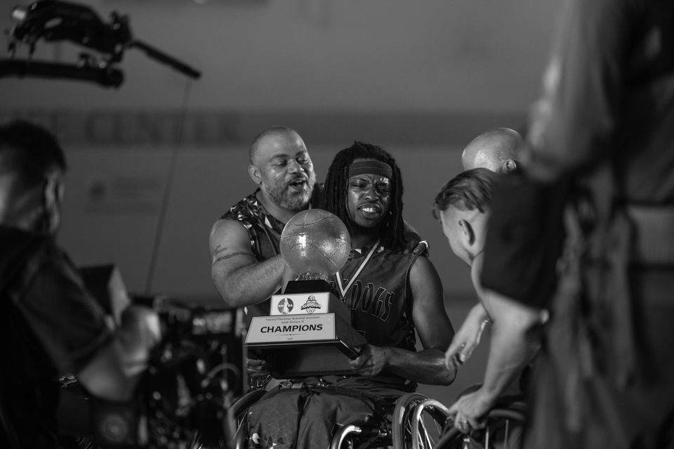 A group of individuals celebrating a victory, with a man in a wheelchair holding a trophy labeled 'CHAMPIONS,' and others gathered around him.