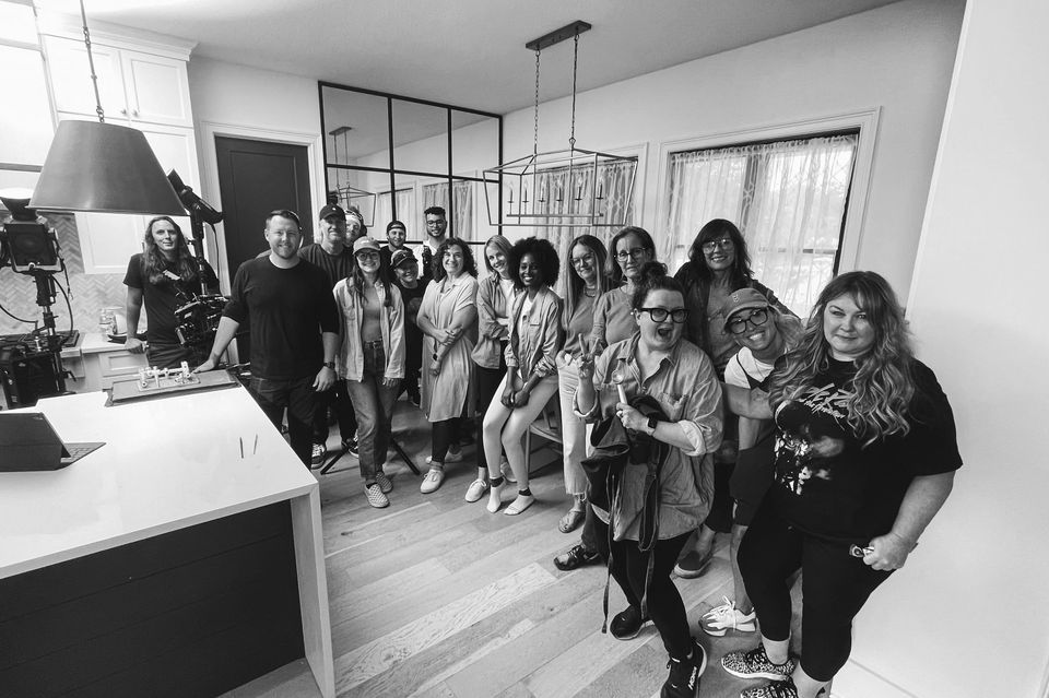 Group of people inside a modern kitchen, smiling and posing for a photo. The room has large windows, curtains, and contemporary lighting fixtures.