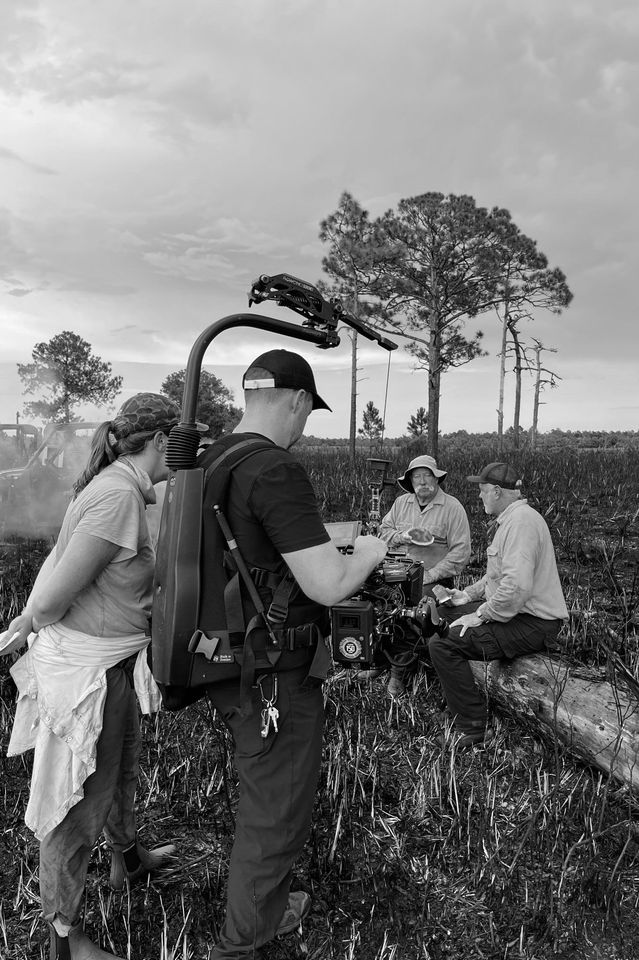 A film crew is recording outdoors in a field, with two men seated on logs and a woman and a man standing nearby, operating a camera. Trees and a cloudy sky are in the background.