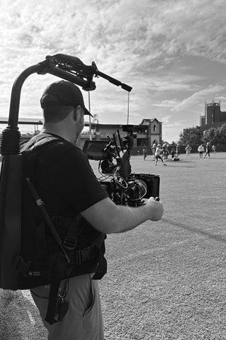 A person operating a camera on a shoulder rig in an outdoor park, with buildings and people in the background.