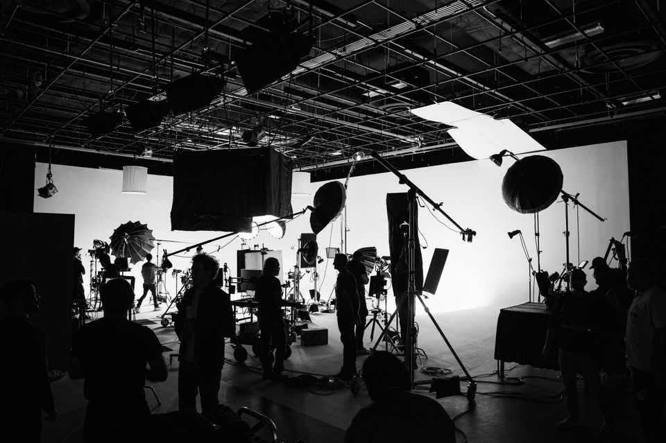 A black and white photo of a film or photography studio with crew members and equipment, including lighting and cameras, silhouetted against a bright background.