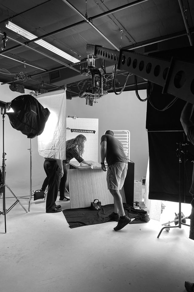 Black and white photo of a photo shoot in a studio with three people working around a table. Equipment includes lighting, a backdrop, and photography gear.