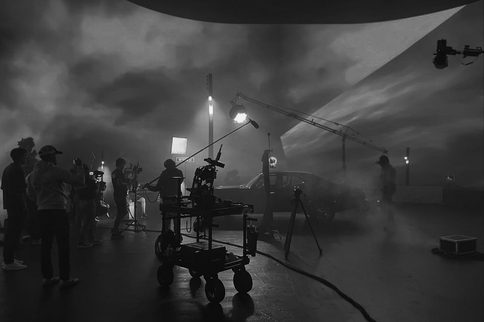 Film crew working on a night shoot with a car, camera equipment, lighting, and crew members under a cloudy sky.