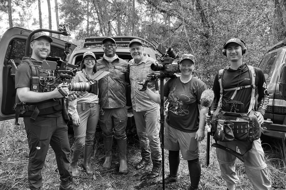 Group of six filmmakers standing outdoors in a forest, some holding cameras and equipment, smiling at the camera in black and white.