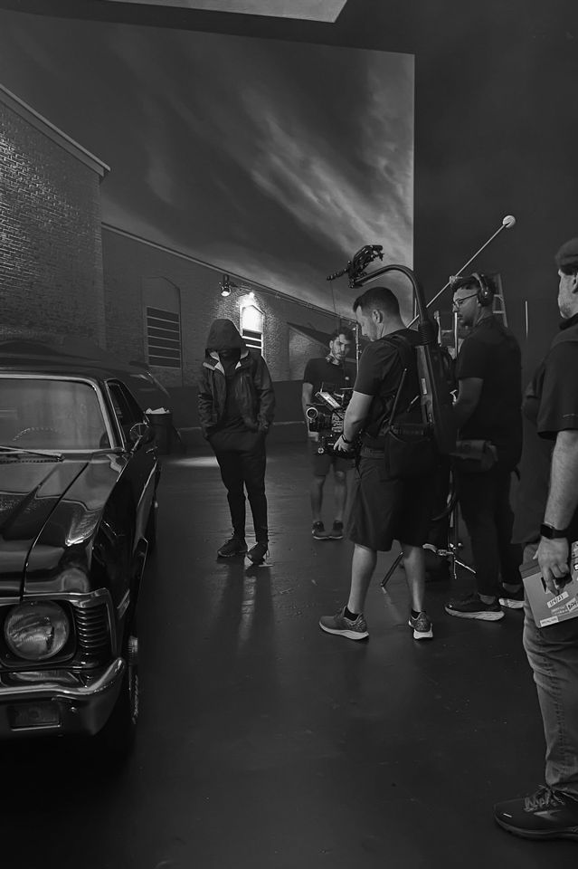 Filming crew on set with a person in a hooded jacket, vintage car, and cloudy sky backdrop.