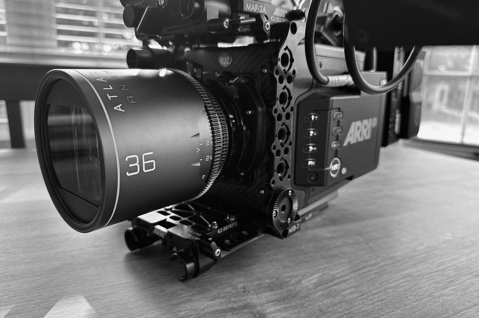 Black and white image of a professional cinema camera with a large lens attached, placed on a wooden surface near a window.