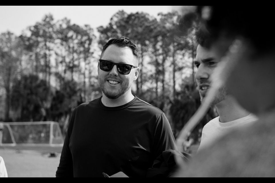 Black and white photo of three men outdoors, one smiling with sunglasses, two with focused expressions, with trees and a sports goal in the background.
