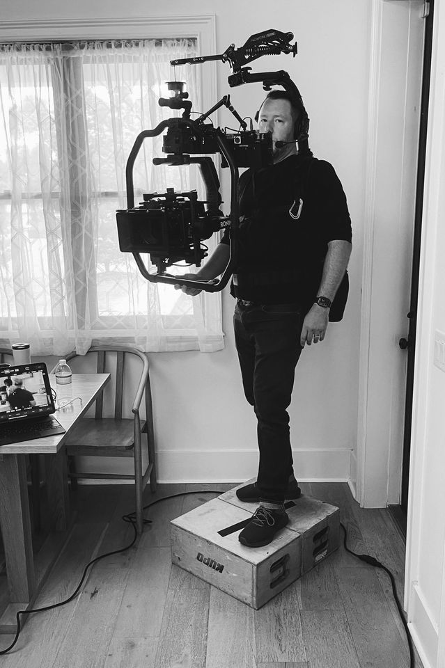 A man wearing a headset and black clothing operating a professional camera mounted on a stabilizer rig, standing on a wooden box in a residential room.