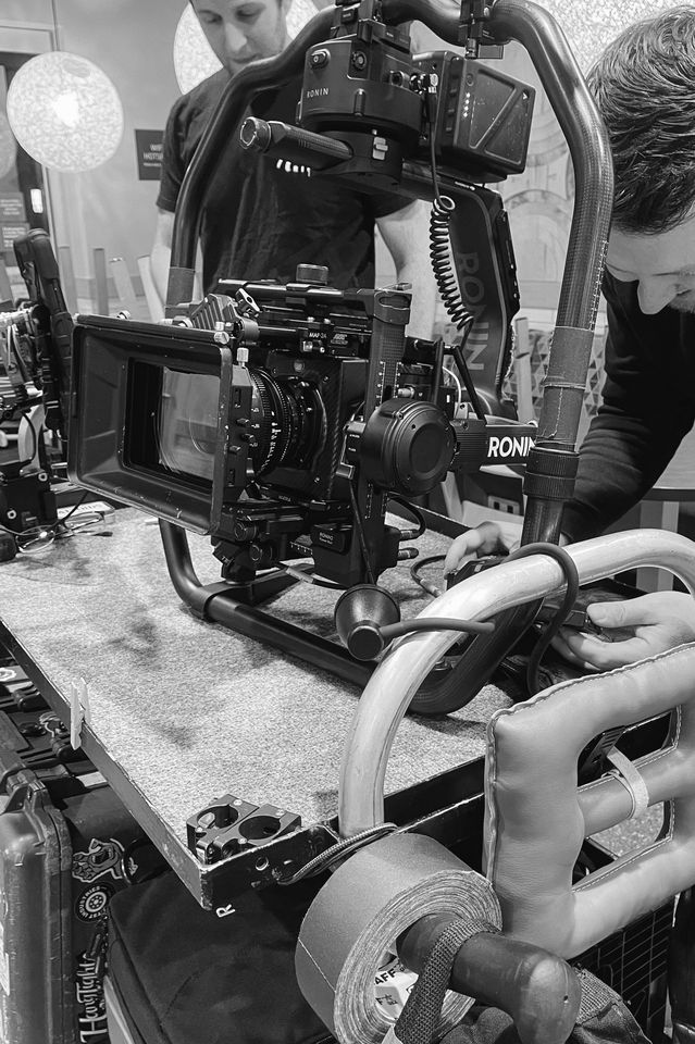 A close-up of a professional film camera mounted on a steadicam rig, with crew members adjusting and preparing the equipment on a studio set.