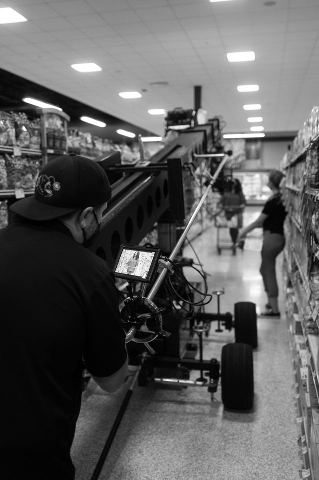 Film crew operating a camera on a dolly in a grocery store aisle, with a woman browsing shelves in the background.
