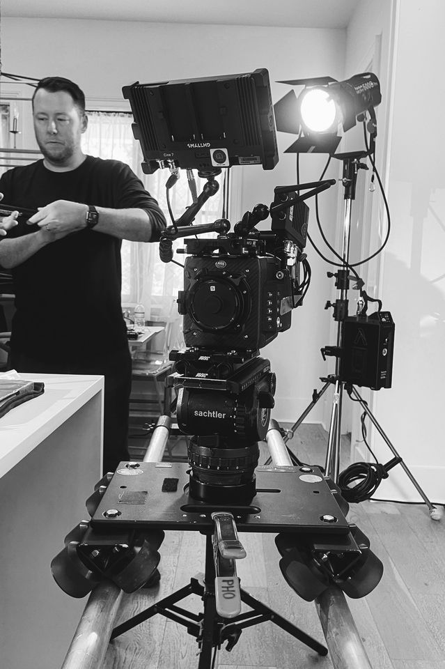 Black and white photo of a professional camera mounted on a sliding tripod with a lighting setup in a room. A man in black shirt is adjusting equipment in the background.