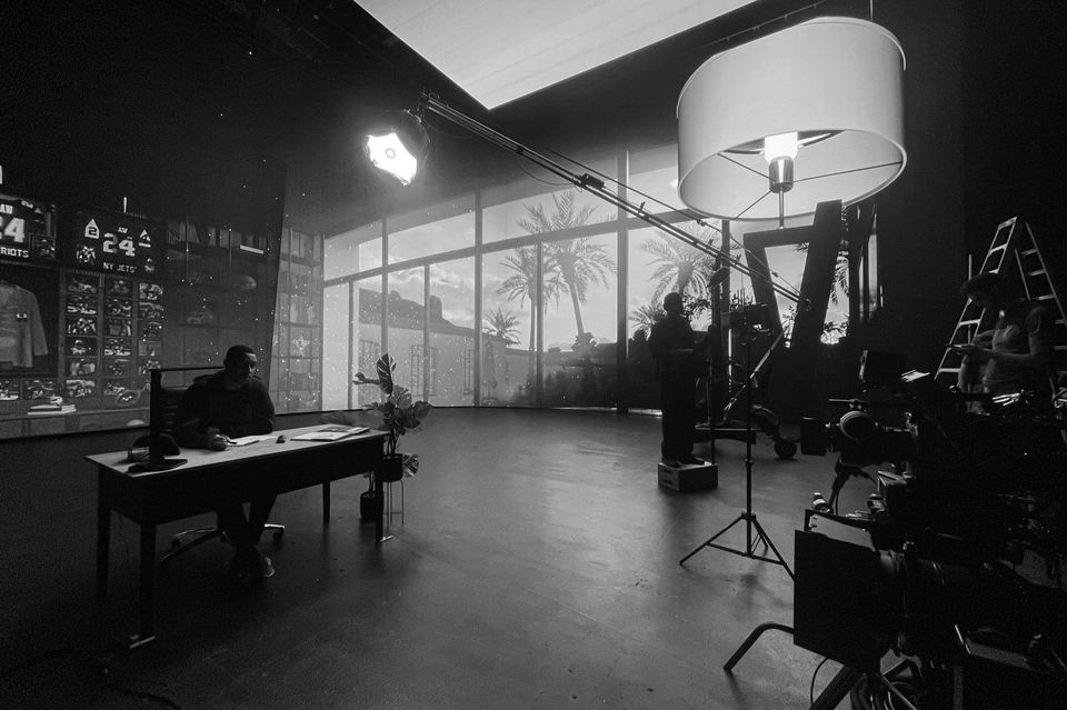 A black-and-white photo of a television or film studio set with large windows showing palm trees and a building outside. Inside, a person is sitting at a desk, surrounded by equipment, with crew members and lighting fixtures set up for filming.