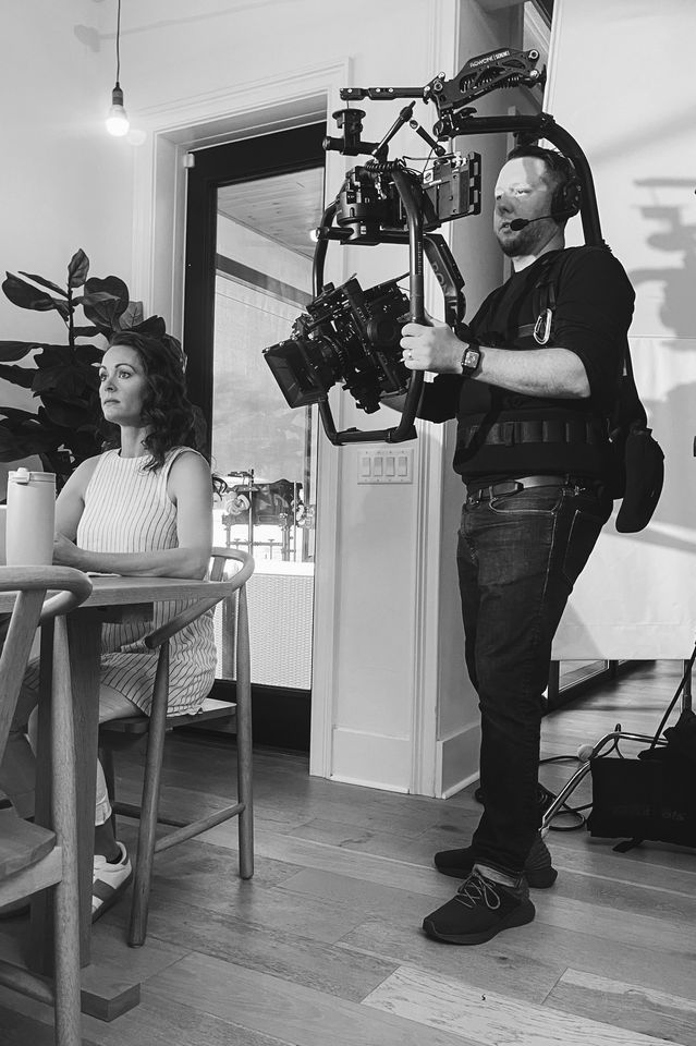 A woman with dark curly hair sitting at a dining table, with a DSLR camera and filming equipment used by a videographer standing nearby, indoors in a home setting.