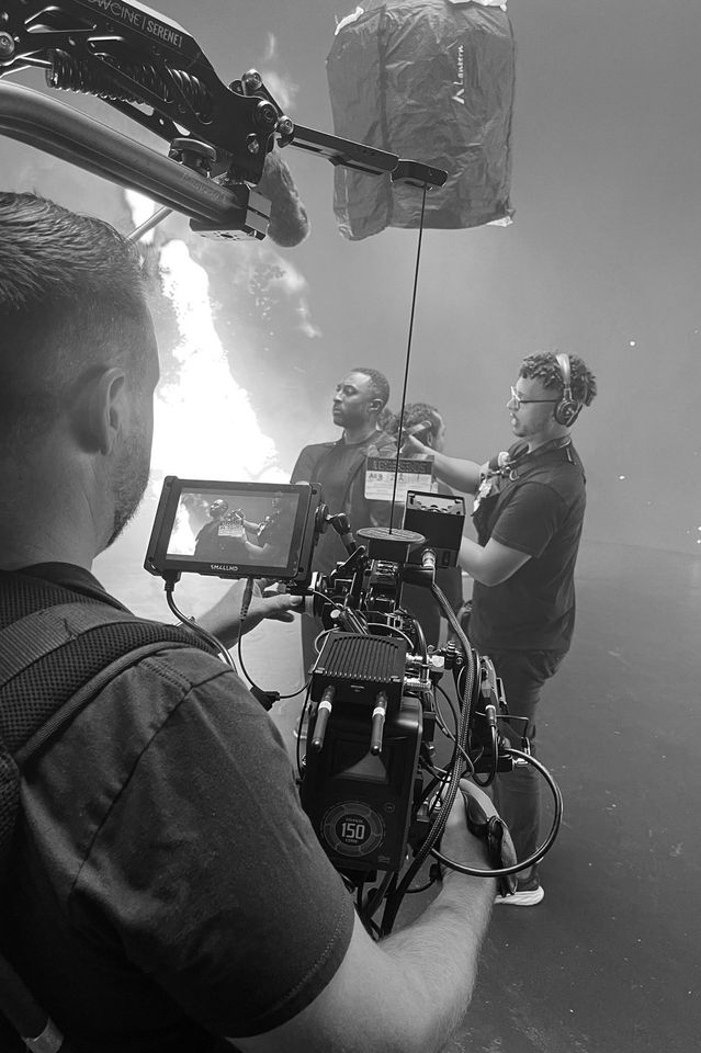 Black and white photo of a film crew filming a scene with a man standing under a large parachute or fabric canopy. The camera operator is focused on the scene, with a monitor showing the shot, while another crew member with headphones is adjusting eq