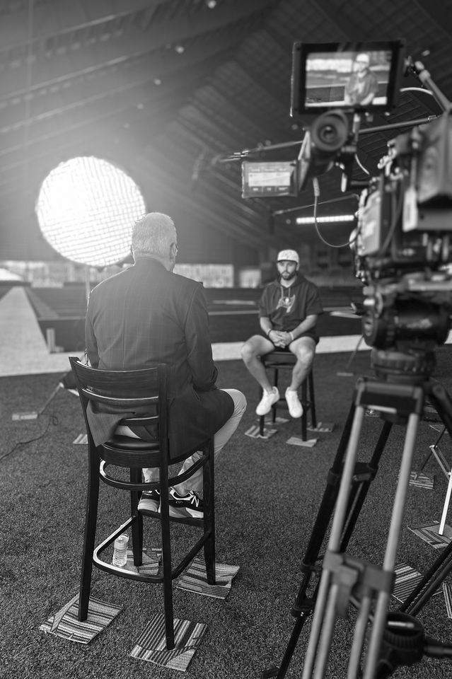 A black and white photo capturing a video interview inside a large indoor sports facility. The interviewer, dressed in a suit, is sitting on a high stool facing a sports player wearing a hoodie, shorts, and a cap, who is also seated. Professional vid