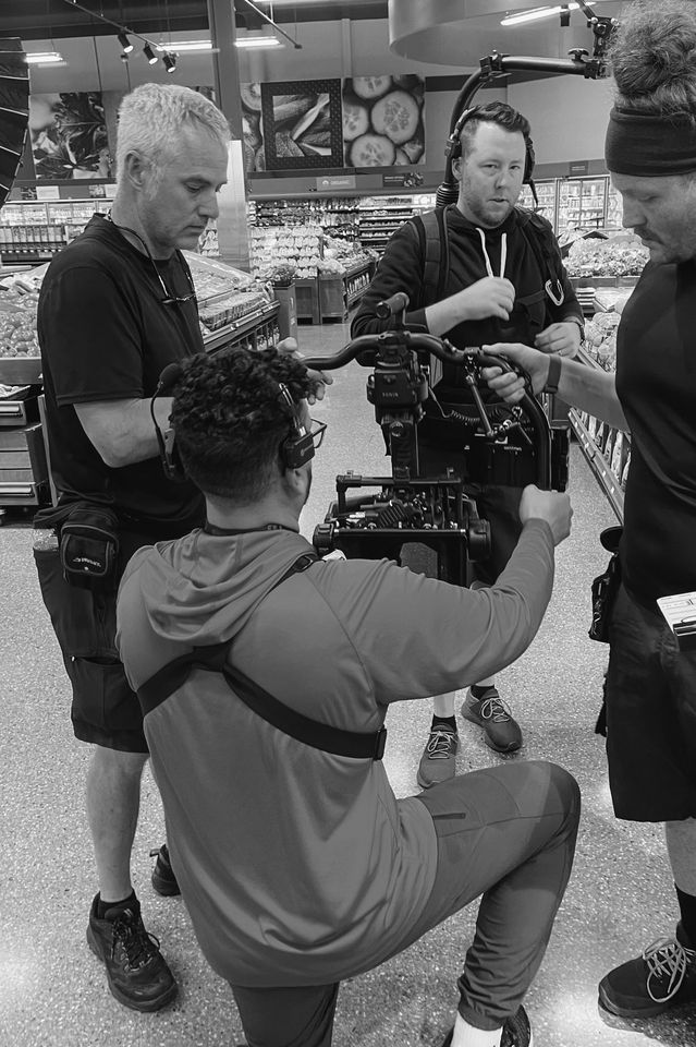 Group of five men filming a scene in a grocery store, with produce and fruit in the background.