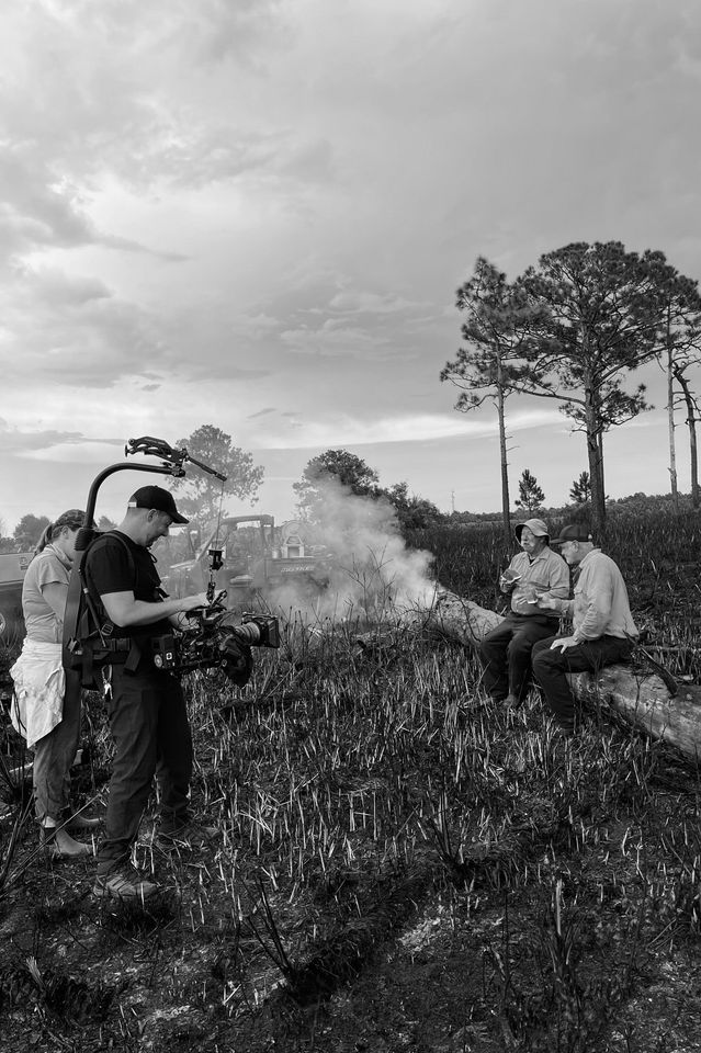 A black and white photo of a small group of people sitting and standing near a smoldering campfire or controlled burn in a grassy area with trees in the background, while a person operates a camera.