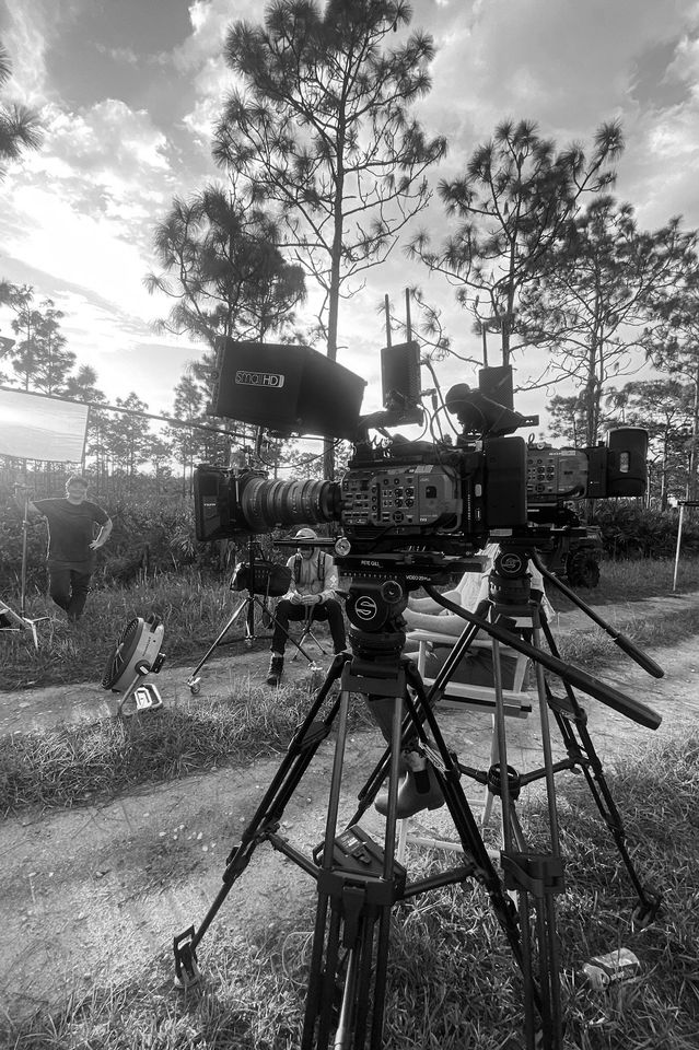 Black and white photo of a film set with a large professional camera mounted on a tripod, with people standing and sitting nearby amid trees, outdoors, under a partly cloudy sky.