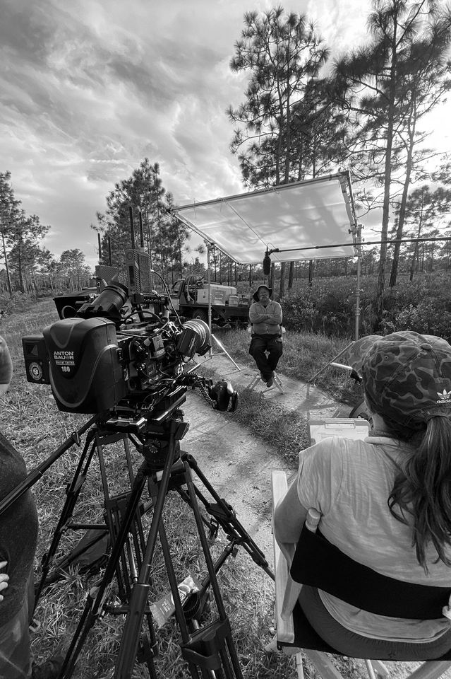 Filming crew setting up outdoor interview scene with a man seated in front of trees and cloudy sky, camera and crew members visible.