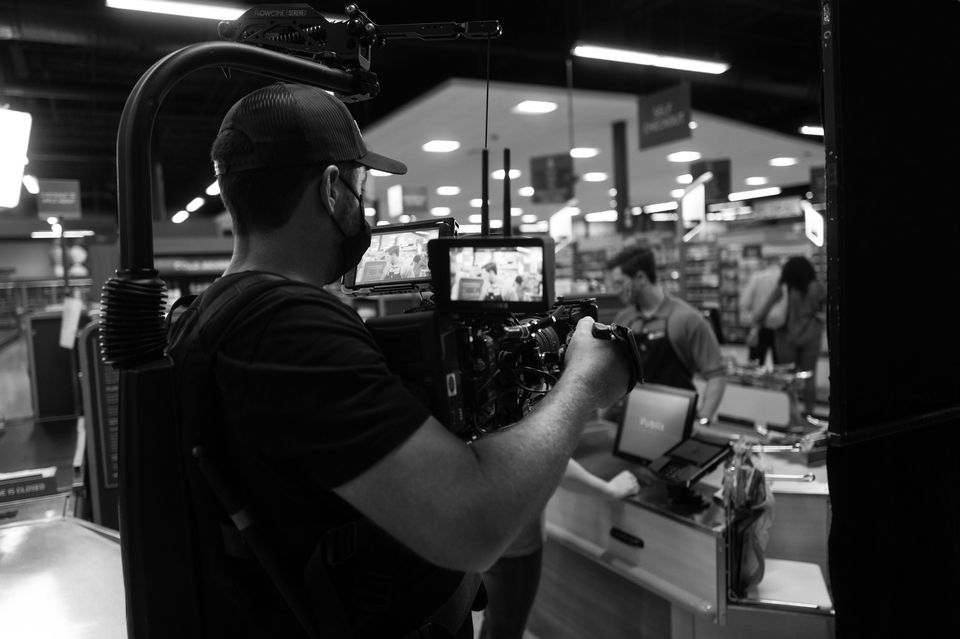 Black-and-white photo of a man wearing a mask, cap, and headphones operating a camera on a film set inside a store. In the background, two people are working at a checkout counter.