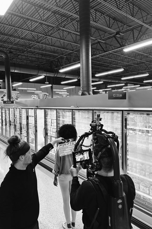 Behind the scenes of a video or film shoot in a grocery store, with a camera operator filming a person holding a clapperboard, and another person assisting the scene.