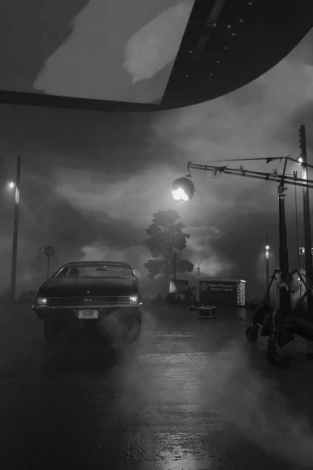 Black and white behind-the-scenes photo of a film set at night, featuring a vintage Chevrolet car, film equipment, large lights, a stop sign, a tree, and a cloudy sky.