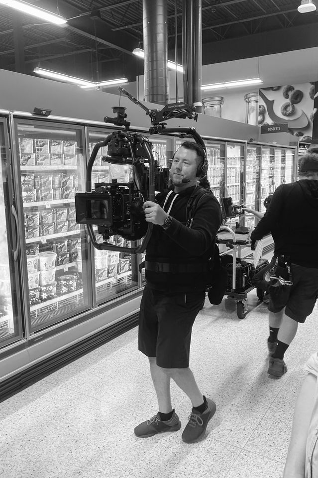 A man is operating professional camera equipment in a grocery store aisle, likely filming or photographing for a video or commercial, with refrigerated shelves of frozen food in the background.