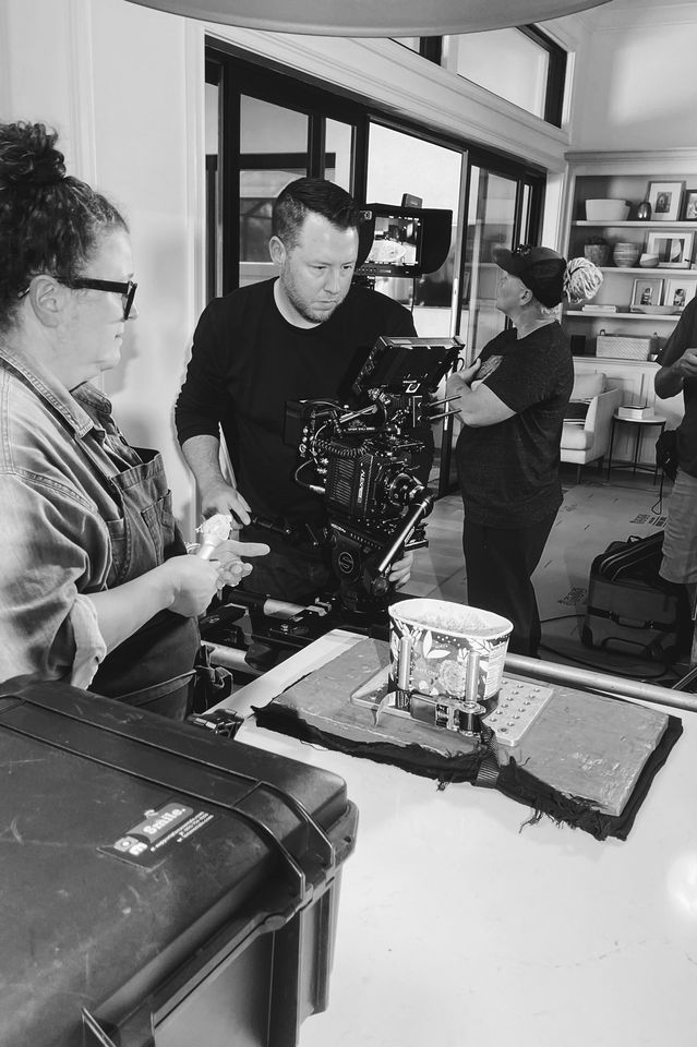 Black and white photo of a film set with crew members preparing for a shot, including a camera operator and a woman holding ice cream, in a room with large windows and shelves with decorative items.