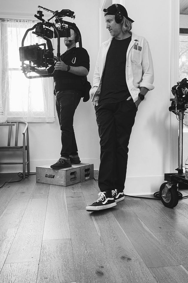 Two crew members on a film set. One is standing, looking down, wearing a baseball cap backwards, a white jacket, and sneakers. The other is holding a camera on a rig, standing on a small wooden box, with headphones on, and a black hoodie.