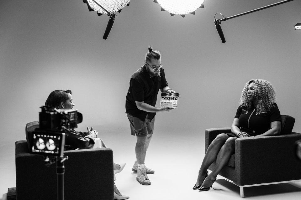 A black and white photo showing a film or TV interview in progress. Two women are seated in armchairs, facing each other. One woman has curly hair and is wearing a dress with a zipper, and the other woman has short hair and glasses. A crew member is 