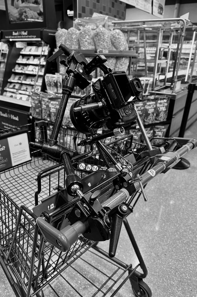 A shopping cart filled with a camera mounted on a tripod in a grocery store aisle.
